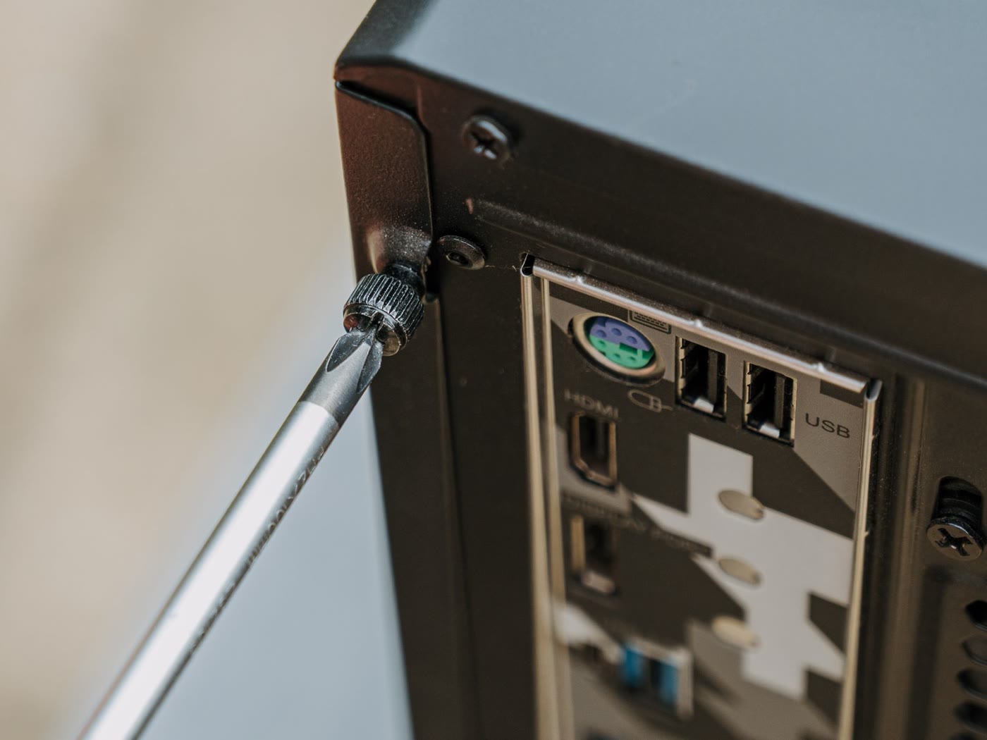 Screwdriver loosening the thumbscrew on the rear corner of a black PC case, above the motherboard rear I/O shield with USB, HDMI and audio ports